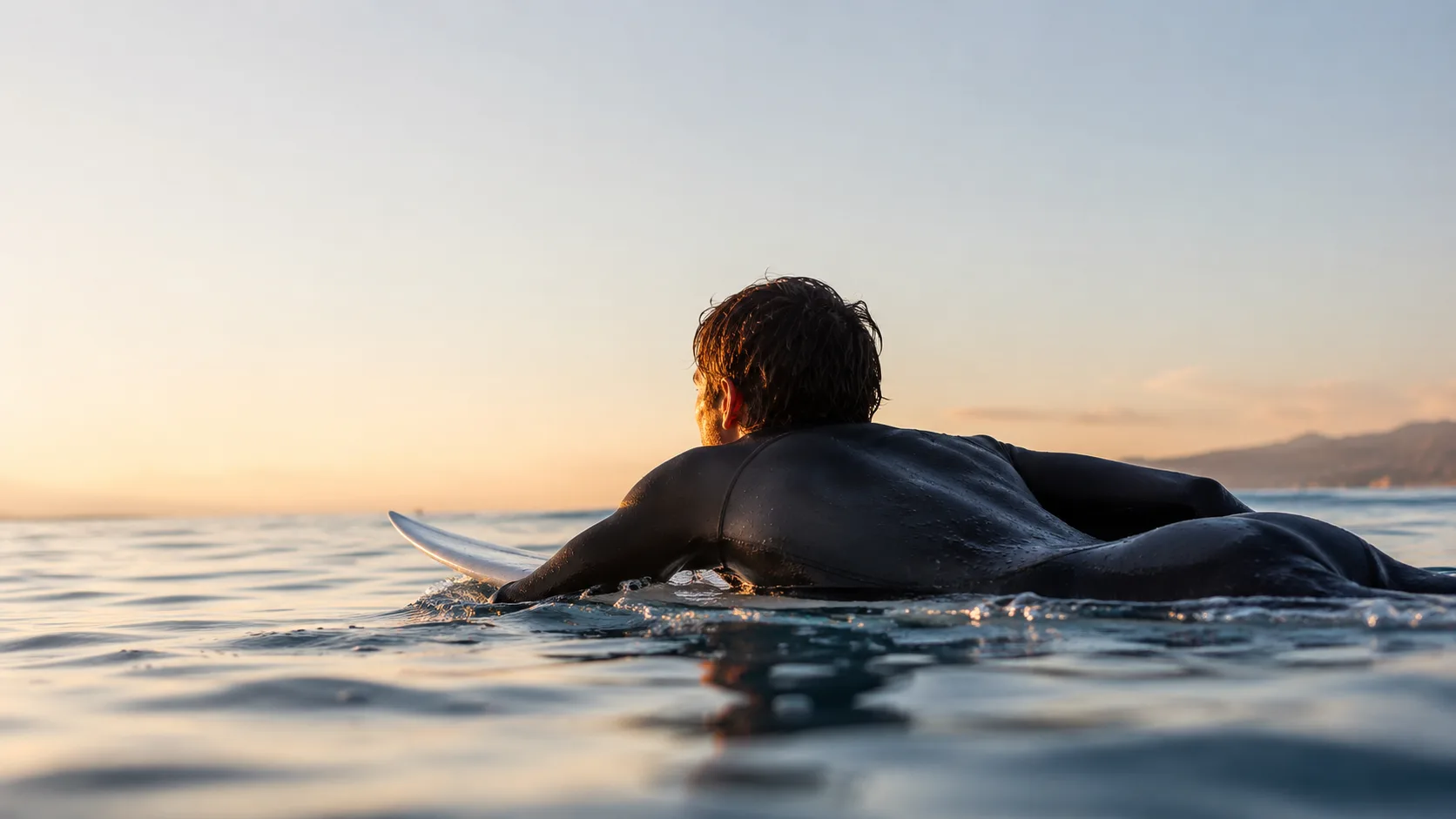 Editorial sports photograph of a surfer paddling out on calm morning water
