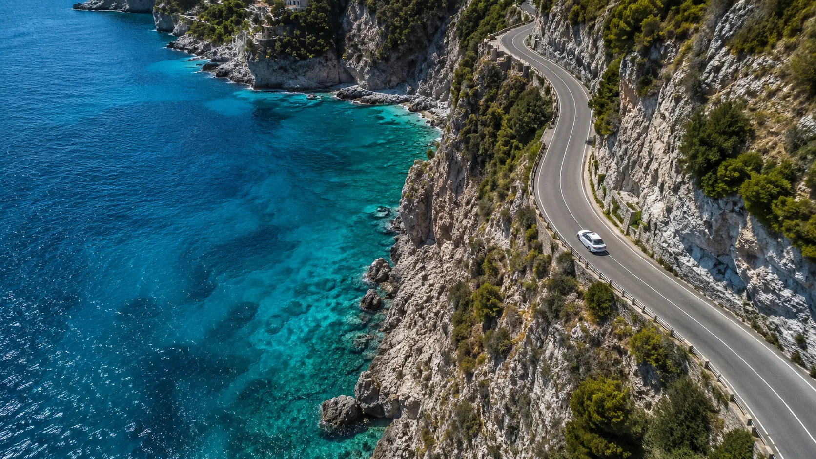 Editorial travel photograph from a drone of a winding coastal road on a cliff with a single white car mid-curve