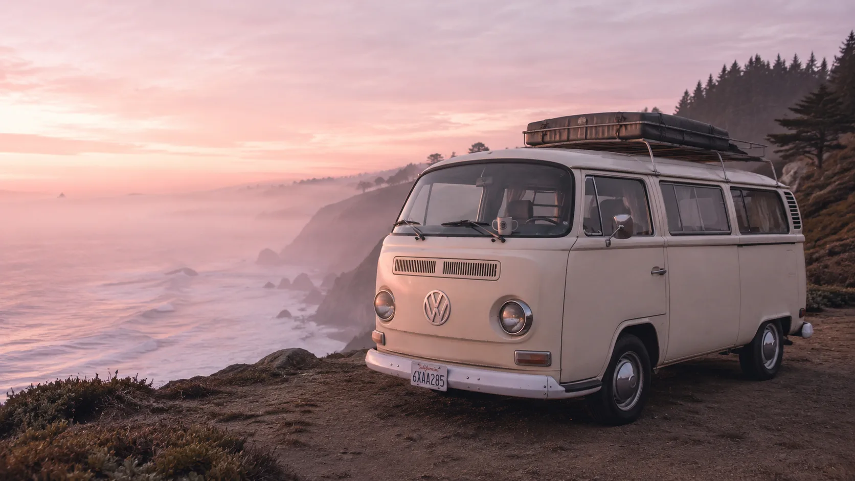 Editorial travel photograph of a vintage cream camper van parked on a coastal cliff at dawn with mist over the ocean