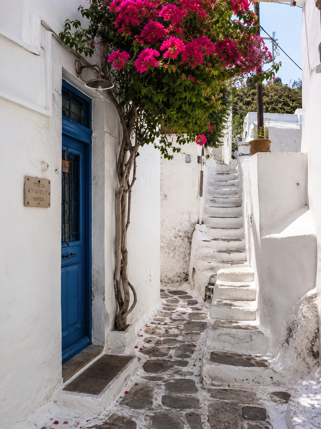 Editorial travel photograph of a narrow whitewashed Greek-island alley with a blue door and bougainvillea spilling over a stair
