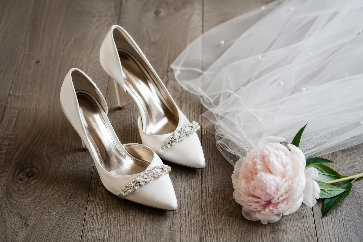 Editorial wedding still life of a pair of ivory satin bridal heels on a wide-plank wood floor with a folded veil and a peony
