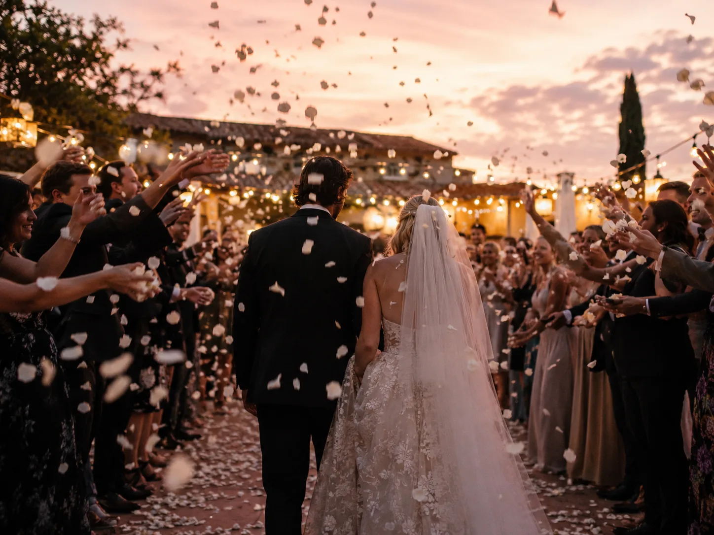 Editorial wedding photograph from behind of a couple walking through a tunnel of guests throwing white petal confetti at dusk