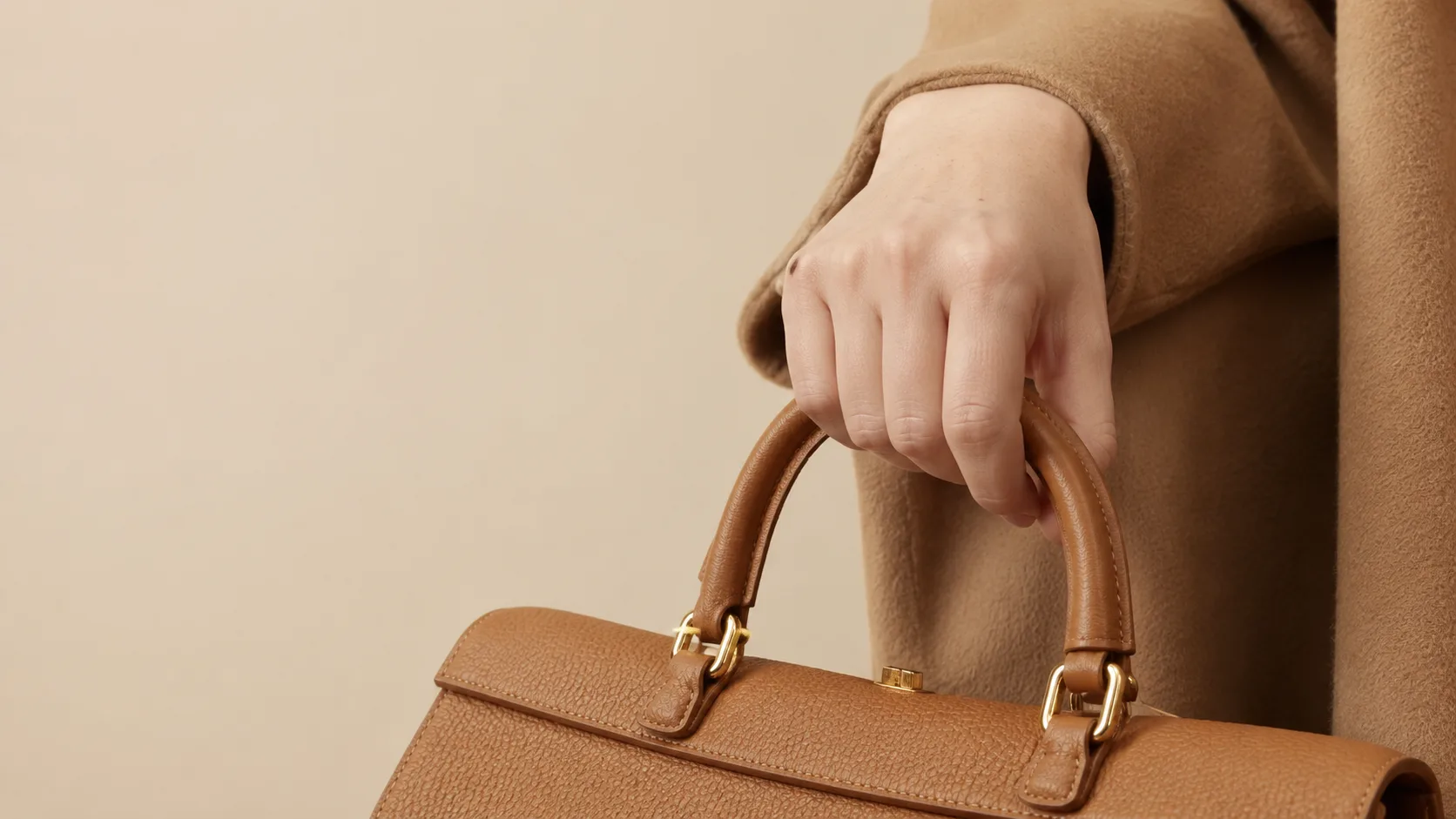 Macro detail of hand emerging from camel coat sleeve holding tan leather handbag
