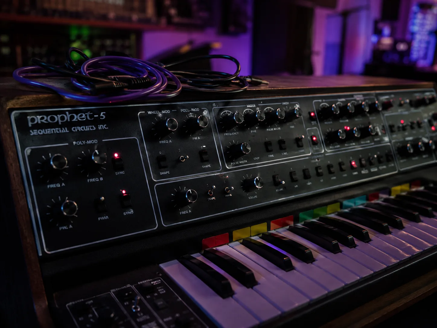 Editorial music close-up of a vintage synthesizer with glowing oscillator LEDs, color-coded keys and coiled patch cables in dim purple ambient light
