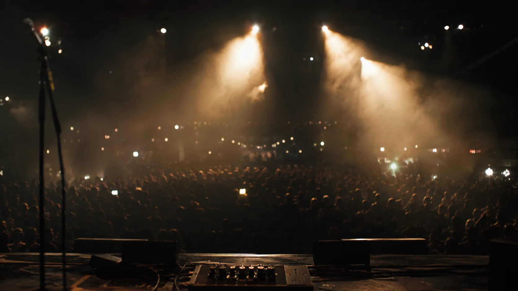 Editorial music photograph from the stage looking out at a dark concert venue with warm spotlight beams cutting through smoke and blurred crowd silhouettes