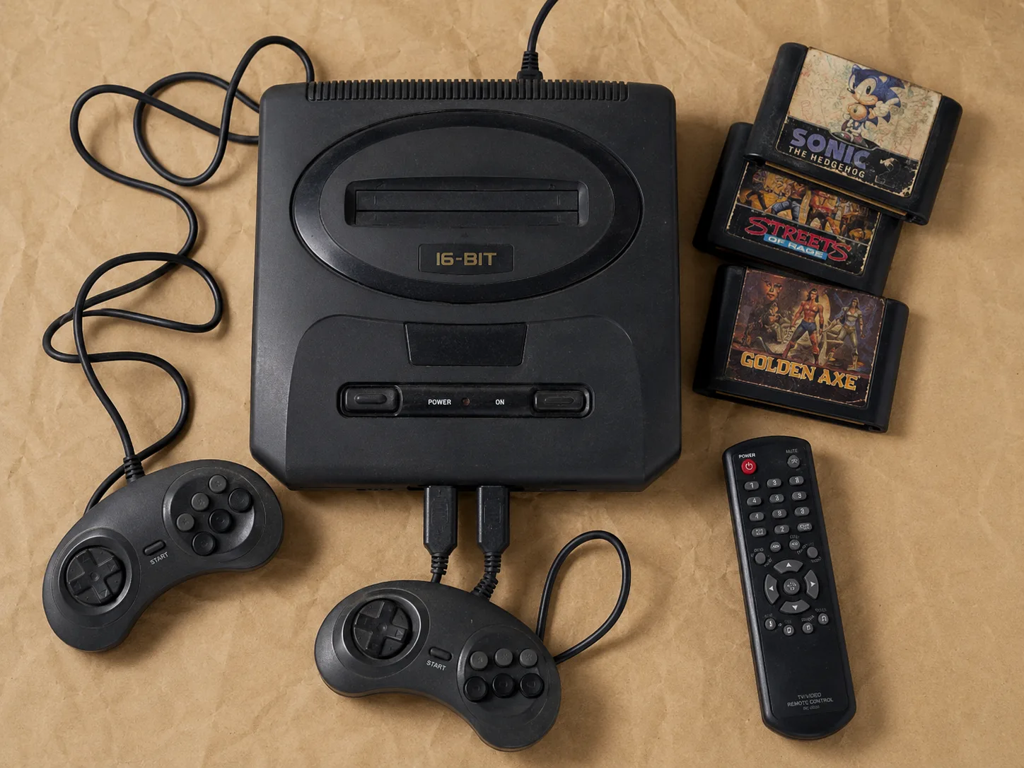 Bodegón cenital de una consola de videojuegos de los 90 con dos mandos con cable y una pila de cartuchos sobre una superficie de papel kraft